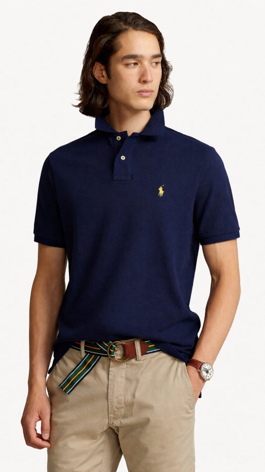 Man wearing a navy blue polo shirt with a small logo, beige pants, and a striped belt on a white background.