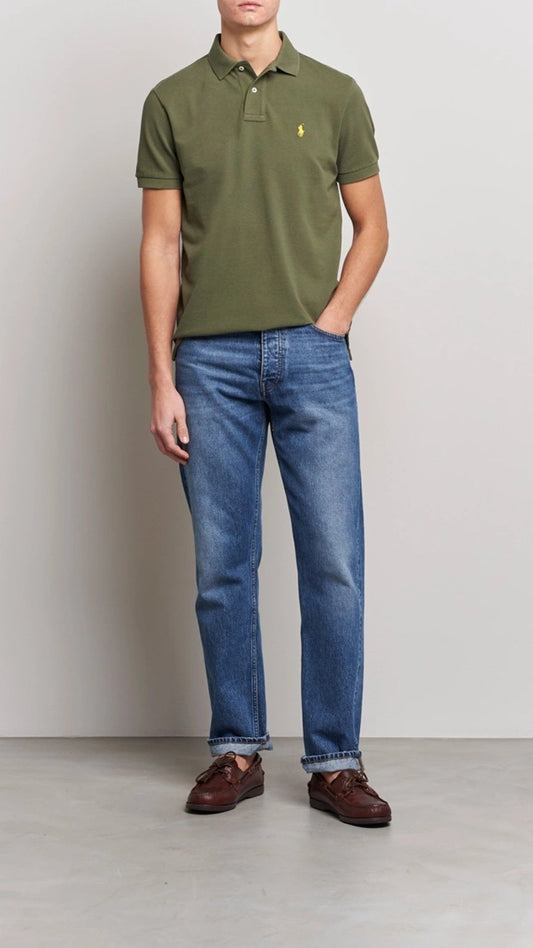 Person wearing a green polo shirt with a logo and blue jeans against a plain background