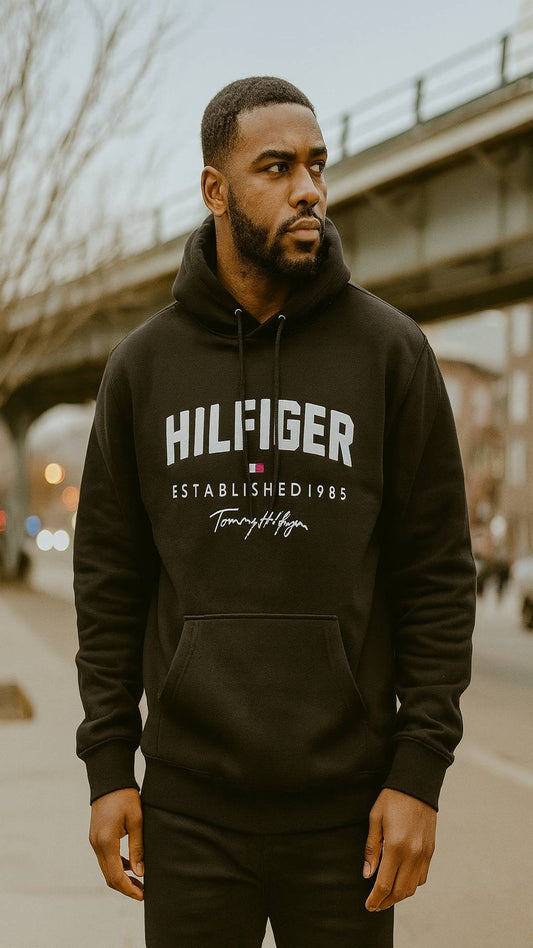 Man wearing a black Tommy Hilfiger hoodie standing outdoors with a bridge in the background.