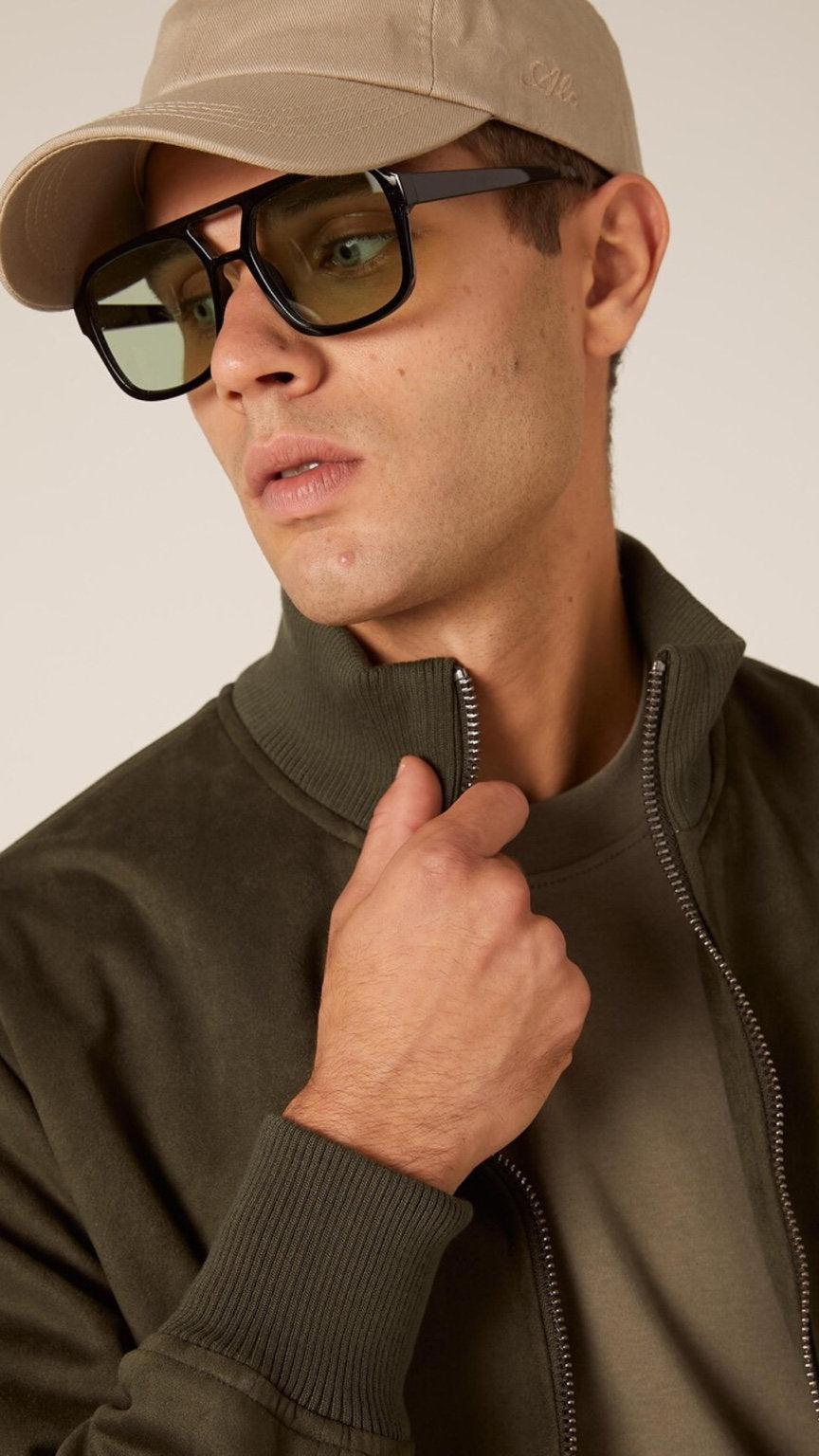 Man wearing a beige cap, sunglasses, and a brown jacket against a neutral background