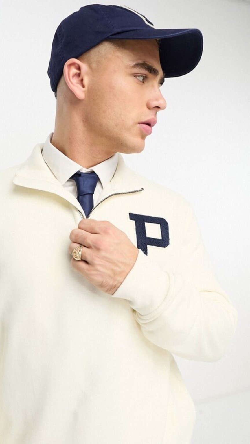 Man wearing a white sweater with a navy 'P' logo, navy cap, and dark tie against a white background