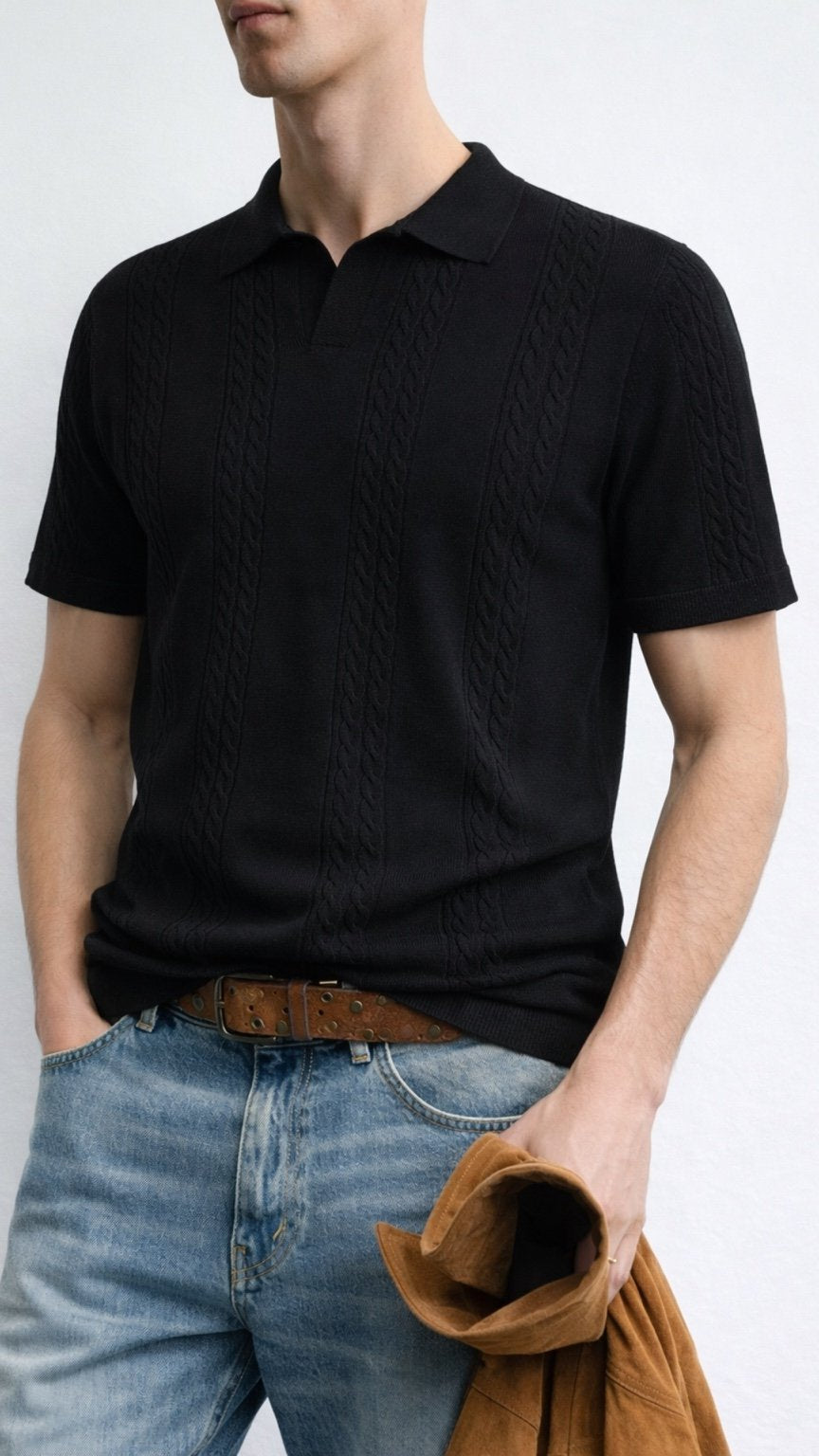 Person wearing a black knitted polo shirt and light blue jeans, holding a brown jacket.