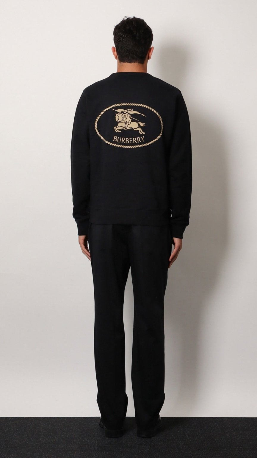 Person wearing a black Burberry sweatshirt with logo on the back against a white wall.
