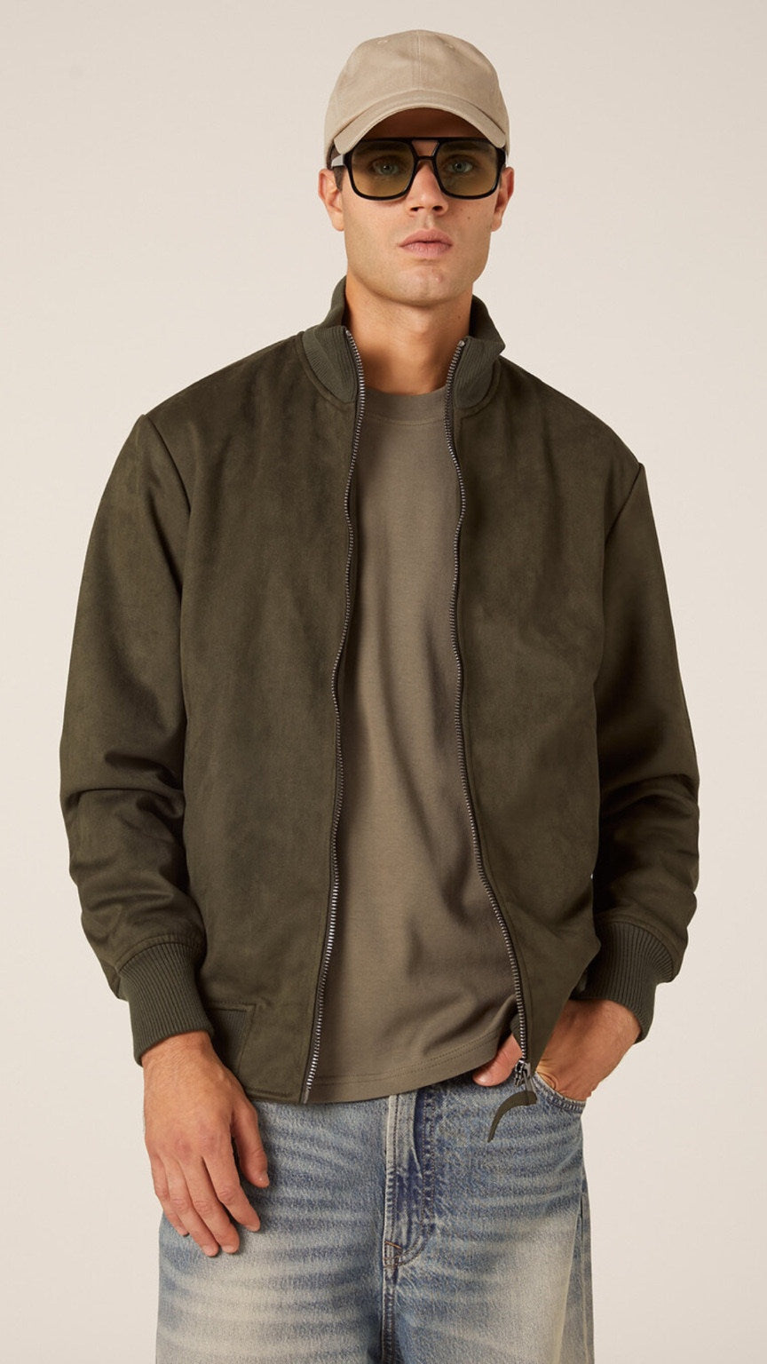 Man wearing a green jacket, gray shirt, blue jeans, beige cap, and sunglasses on a white background