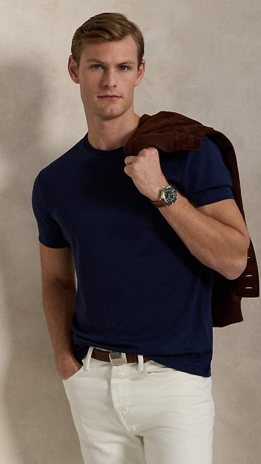 Man wearing a navy blue t-shirt and white pants, holding a brown jacket against a beige background