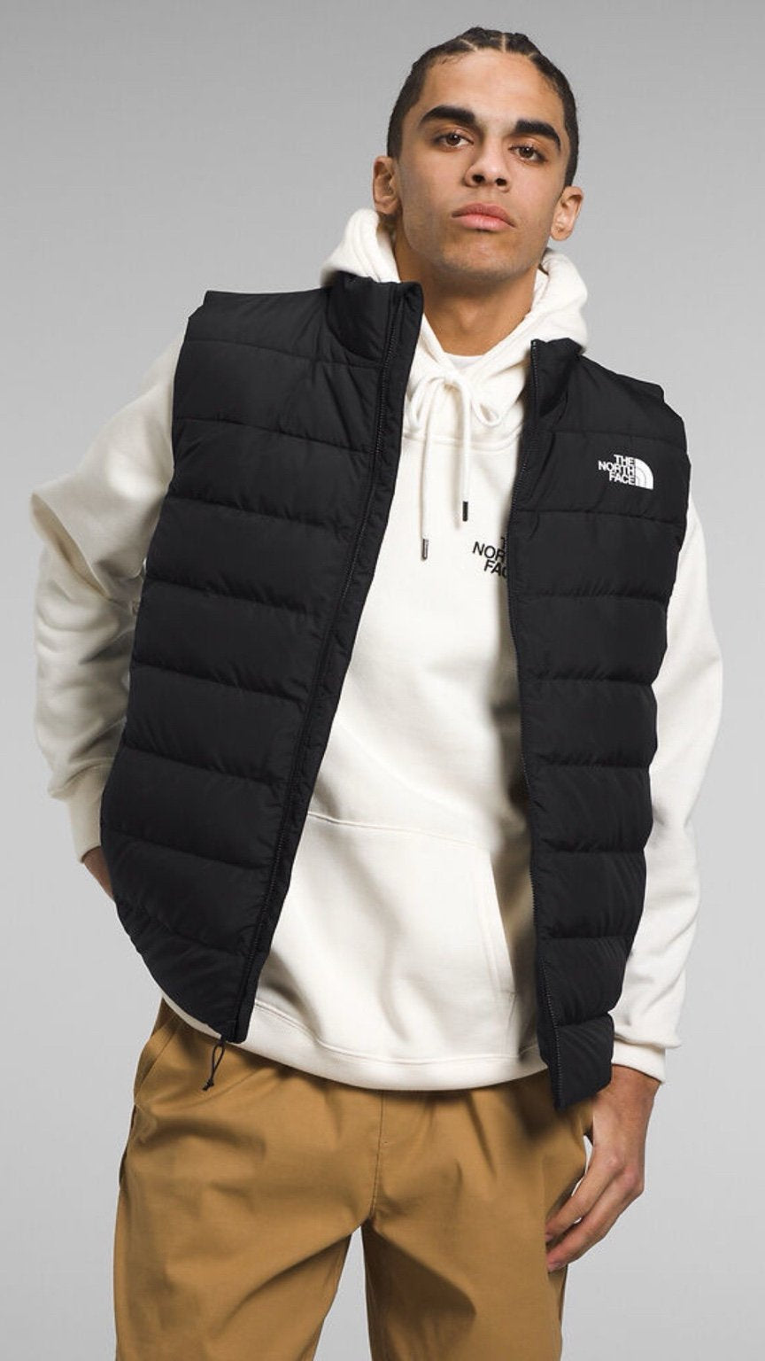 Person wearing a black puffer vest over a white hoodie with a brand logo on a gray background